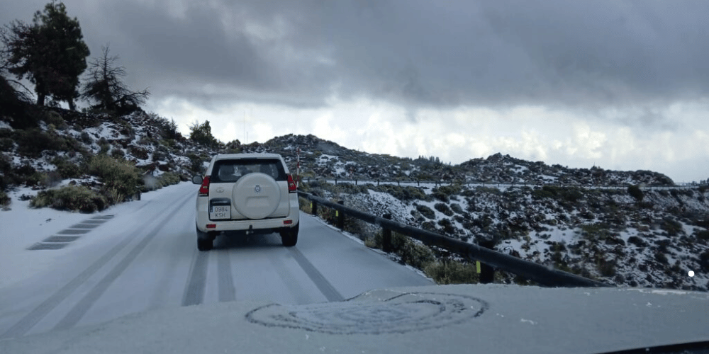 Pico del Teide nevado, con vehículos circulando por la carretera TF-21 bajo un cielo nublado.